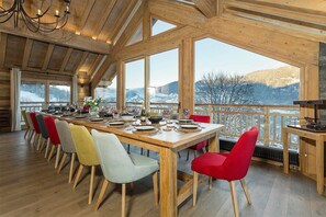 Ein Essbereich mit einem langen Holztisch, gedeckt für eine Mahlzeit, große Fenster mit Blick auf schneebedeckte Berge und eine Holzzielendecke mit freiliegenden Balken.
