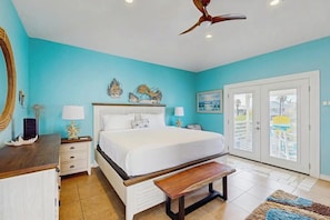 A bedroom with a bed, bedside tables, a ceiling fan, a bench, a window with a view, and a door.