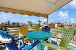 A covered outdoor area with a round table and colorful Adirondack chairs, overlooking a residential neighborhood with houses and a clear sky.