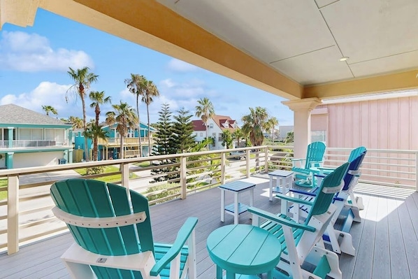 A covered outdoor deck with turquoise chairs and tables, a railing, and a view of palm trees and buildings.