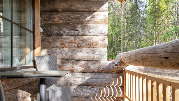 A wooden deck with a table and chairs, a log wall, and a view of trees.