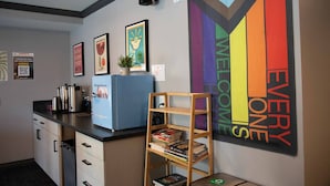 A modern kitchen with a coffee machine, a kettle, and a small refrigerator. There is a wooden shelf with books and a potted plant. A colorful welcome sign is on the wall. - Samesun San Francisco - Hostel (San Francisco)