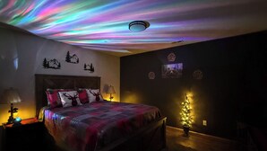 Un dormitori amb un llit, una tauleta de nit, una làmpada penjada a la paret, un projector de sostre i un petit arbre de Nadal.