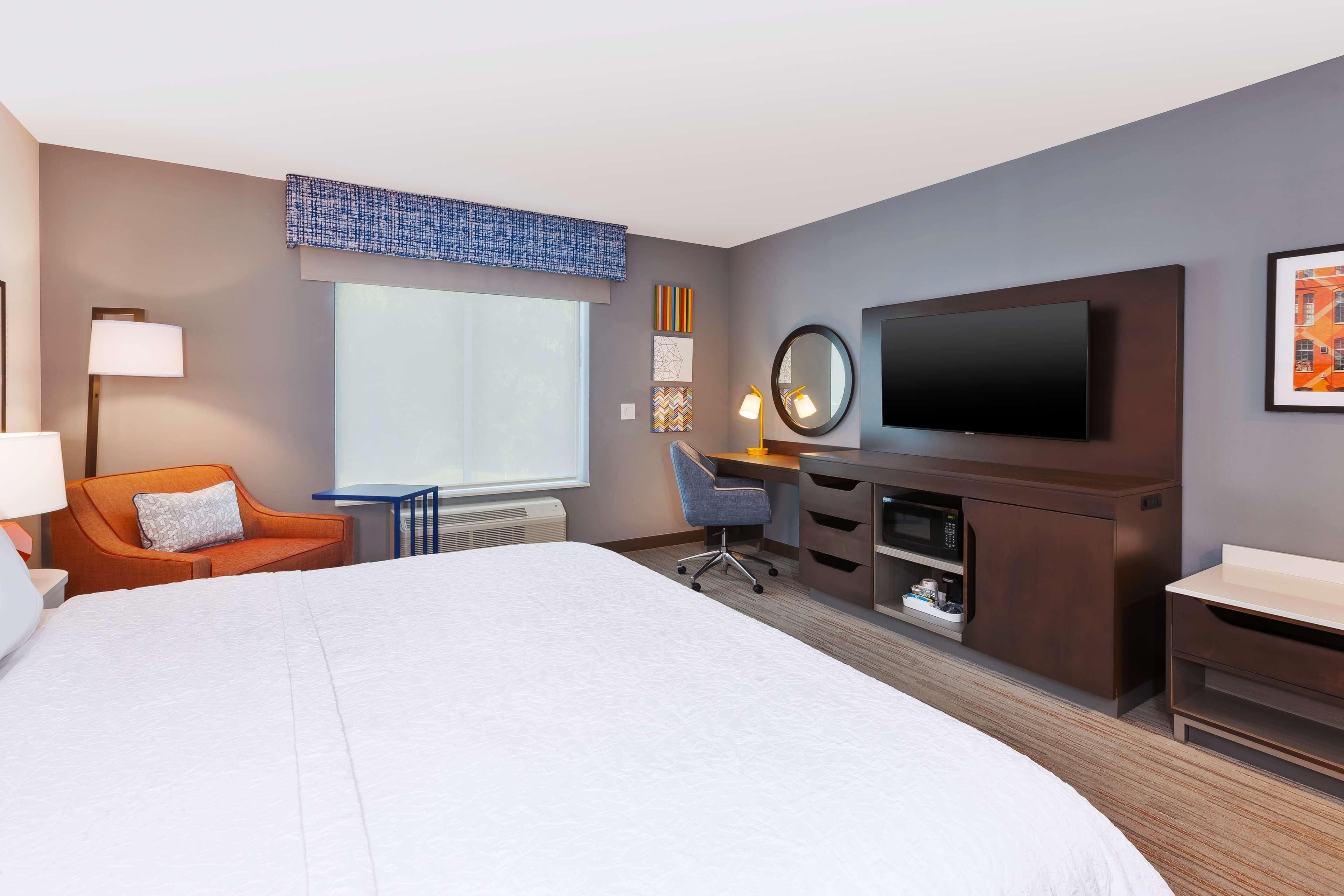 A hotel room with a large bed, a desk with a chair, a television, and a window with blinds.