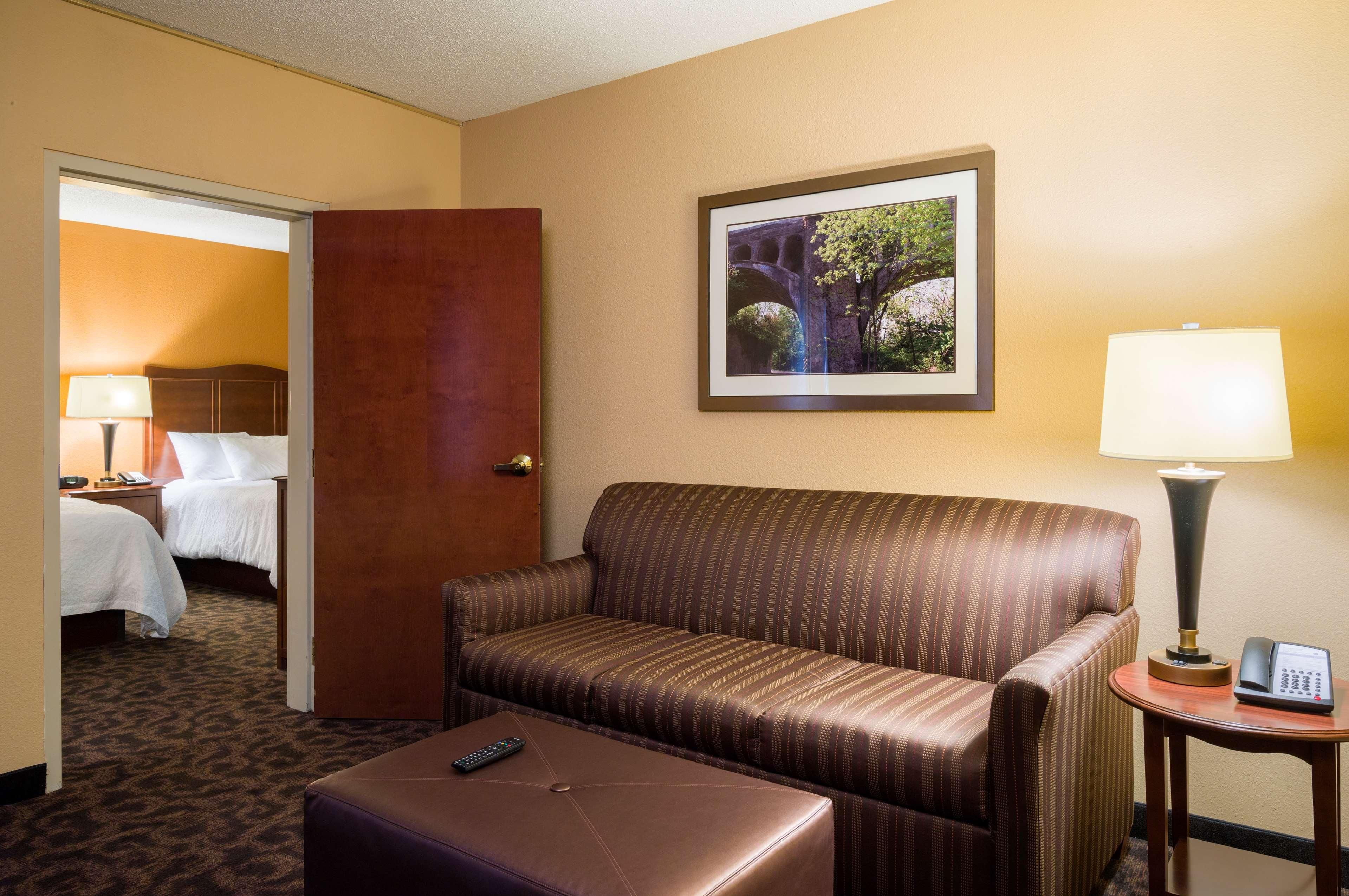 A hotel room with a striped sofa, a small table with a lamp, a remote control on a ottoman, and a framed picture on the wall.