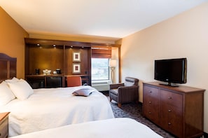 A hotel room with two beds, a TV on a wooden dresser, a chair, and a desk with a lamp.
