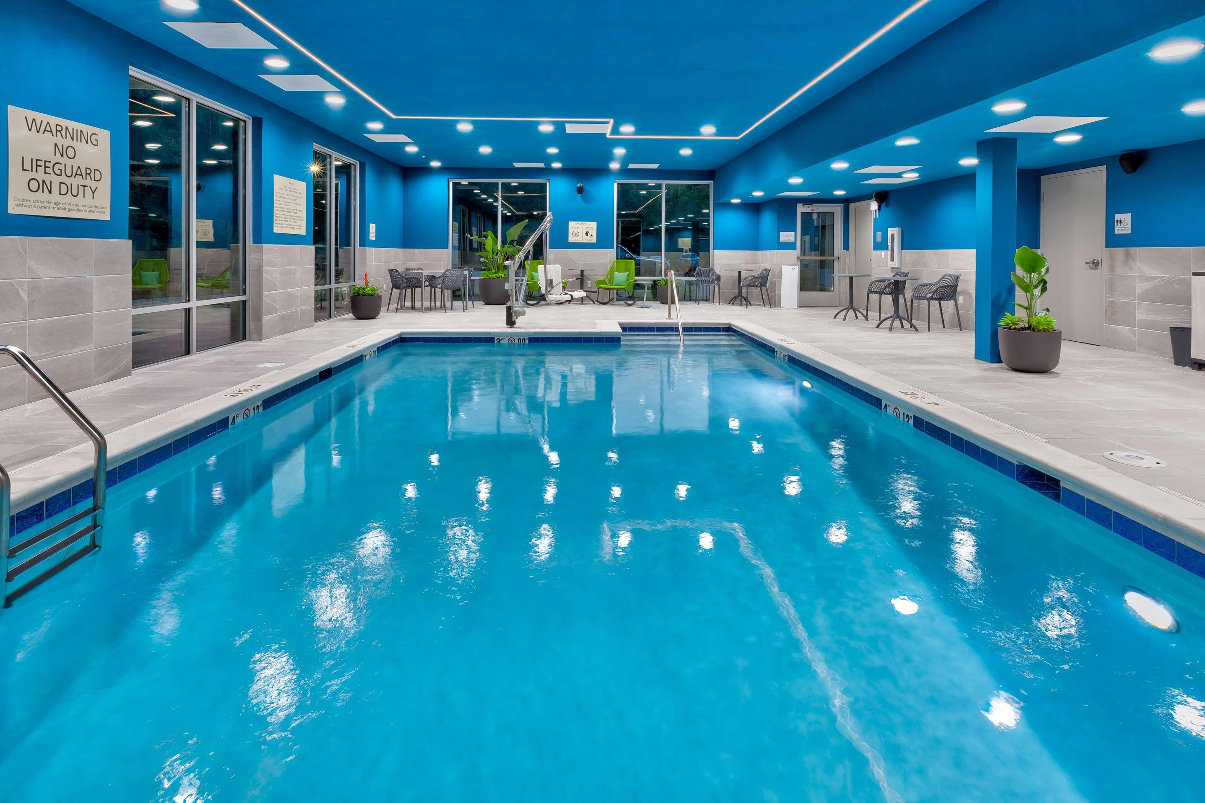 Une piscine aux murs et au plafond recouverts de carreaux bleus, entourée de chaises et de tables, et un panneau indiquant la présence d’un sauveteur sur le mur.