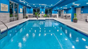 Une piscine aux murs et au plafond recouverts de carreaux bleus, entourée de chaises et de tables, et un panneau indiquant la présence d’un sauveteur sur le mur.