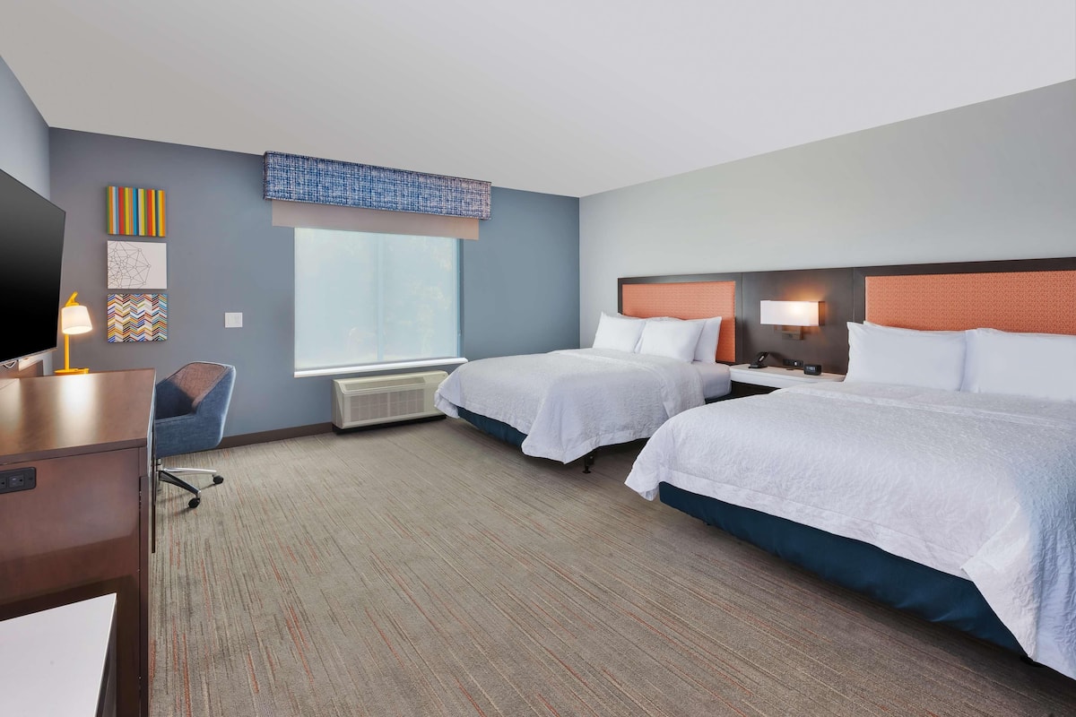 A hotel room with two beds, a desk, a chair, a TV, and artwork on the wall.
