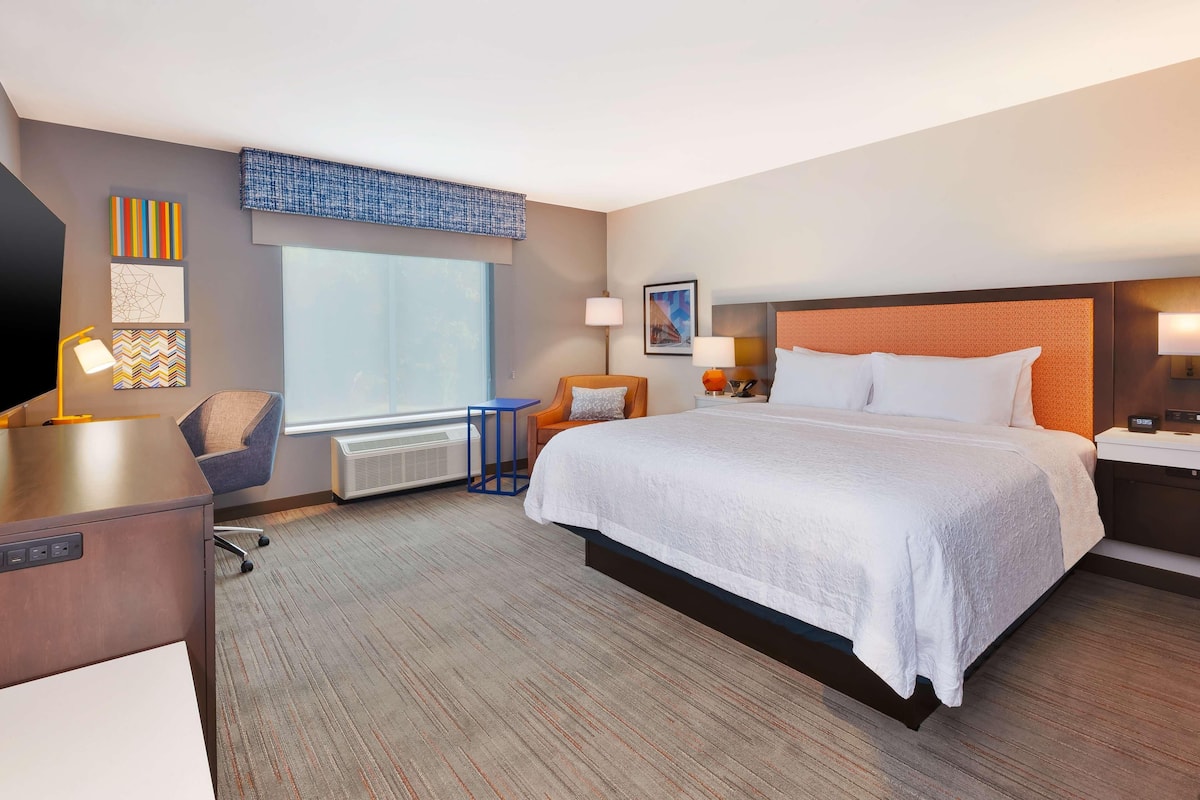 A hotel room with a large bed, a desk, a chair, a window, and artwork on the walls.