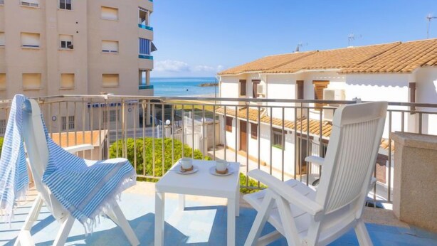 A balcony with white chairs and a table, overlooking a beach and a building with a tiled roof. - Blue Sea by Interhome (Cunit)