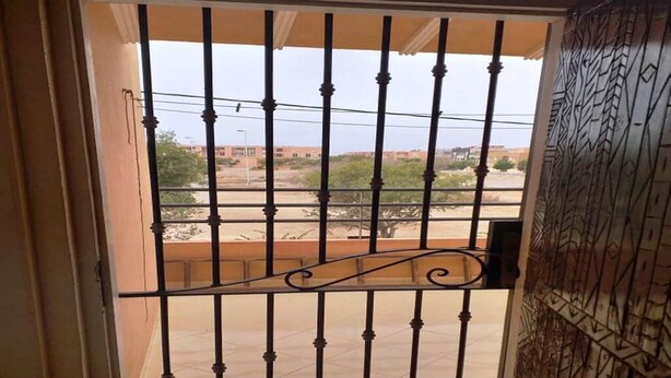 Un balcón con una reja metálica, piso de baldosas y vista a edificios distantes.