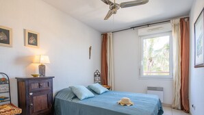 Een slaapkamer met een bed, een nachtkastje, een lamp, een raam met gordijnen en een plafondventilator.