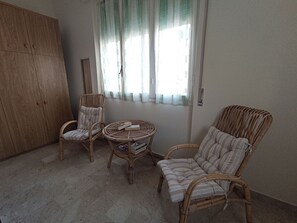 A room with a wooden wardrobe, two wicker chairs, a small round table with books, and a window with sheer curtains.