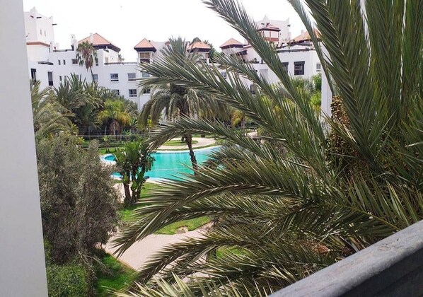Vista de una piscina rodeada de palmeras y un edificio con fachada blanca y techos rojos.