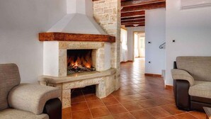 A stone fireplace with a fire, wooden beams, and a tiled floor in a room with two upholstered chairs. - Villa with pool, sauna, sun terrace and WIFI (Golaš)
