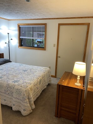Une chambre à coucher avec un lit, une fenêtre avec des stores, une porte et une table de chevet avec une lampe.