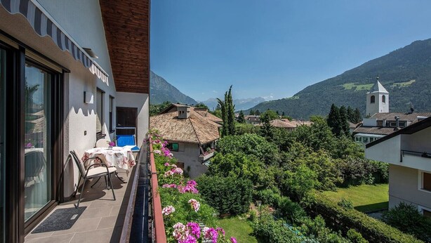 A balcony with a table and chairs, a view of a village with houses and a church, and mountains in the background. - Apartment "Schnitzer - Enzian" with Mountain View, Wi-Fi, Balcony & Garden (Rabland/Partschins)