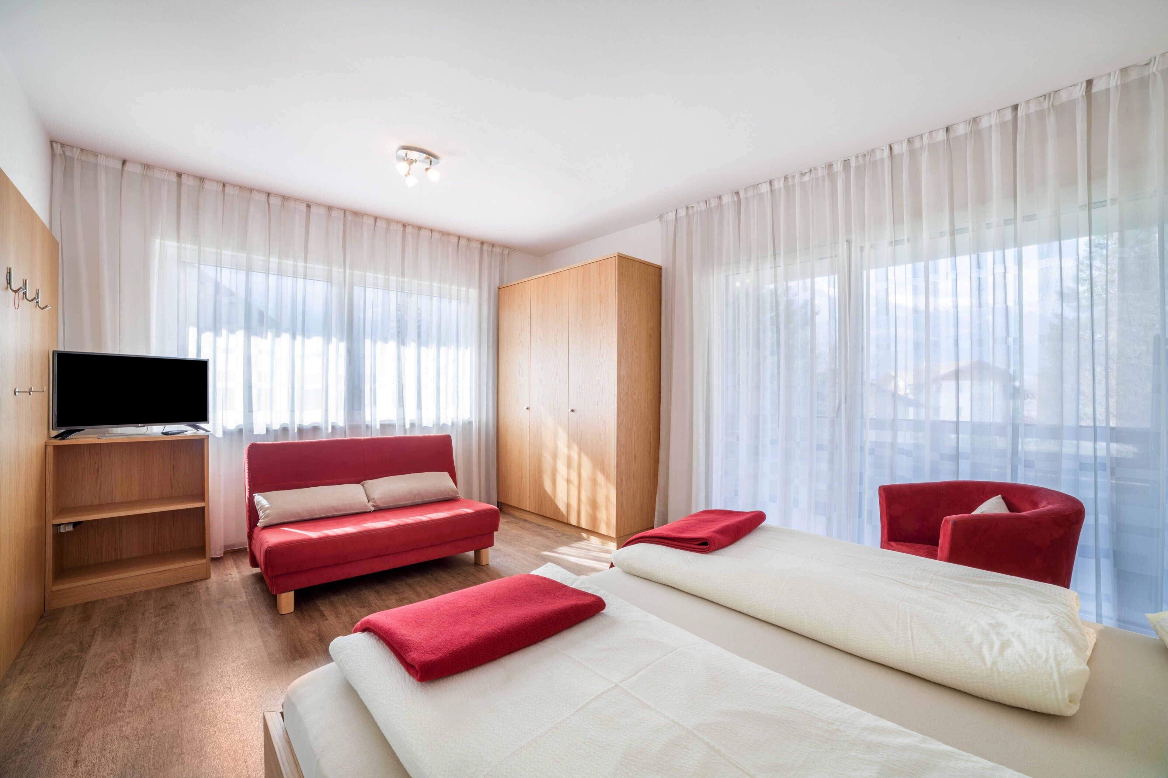 Una camera d'albergo con una TV, un divano rosso, un letto, una poltrona rossa e un armadio.