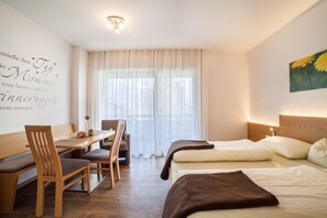 A hotel room with a bed, a desk, chairs, and a window with sheer curtains.