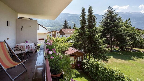 Un balcón con mesa y sillas, con vistas a un jardín con árboles y una montaña.