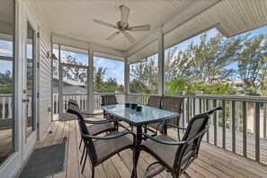 Un espace extérieur abrité, équipé d’une table et de chaises, d’un ventilateur de plafond, et offrant une vue sur des arbres et une maison.