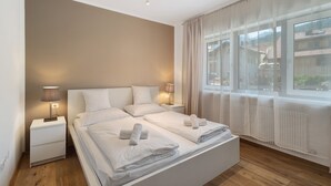 A hotel room with a large bed, two bedside tables, a window with curtains, and a view of buildings outside. - Premium Apartment Hofer Sandra – Fast Internet, Free Parking (Niederdorf)