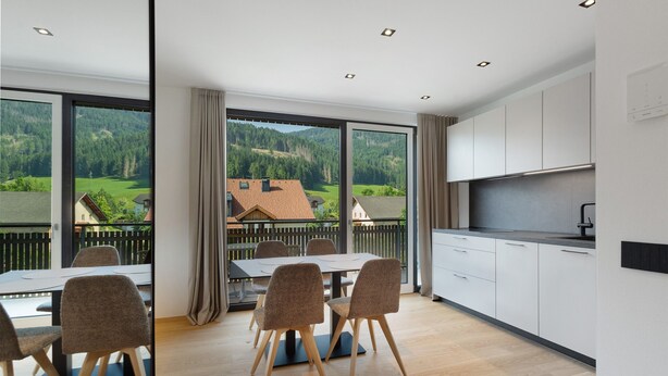 A modern kitchen with white cabinets, a dining area with a table and chairs, and large windows offering a view of a mountainous landscape. - Premium Apartment Hofer Heidi – Fast Internet, Free Parking (Niederdorf)