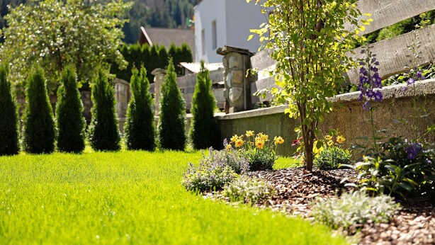 En välskött trädgård med ett stort utbud av växter och blommor, en stenmur och en byggnad i bakgrunden.