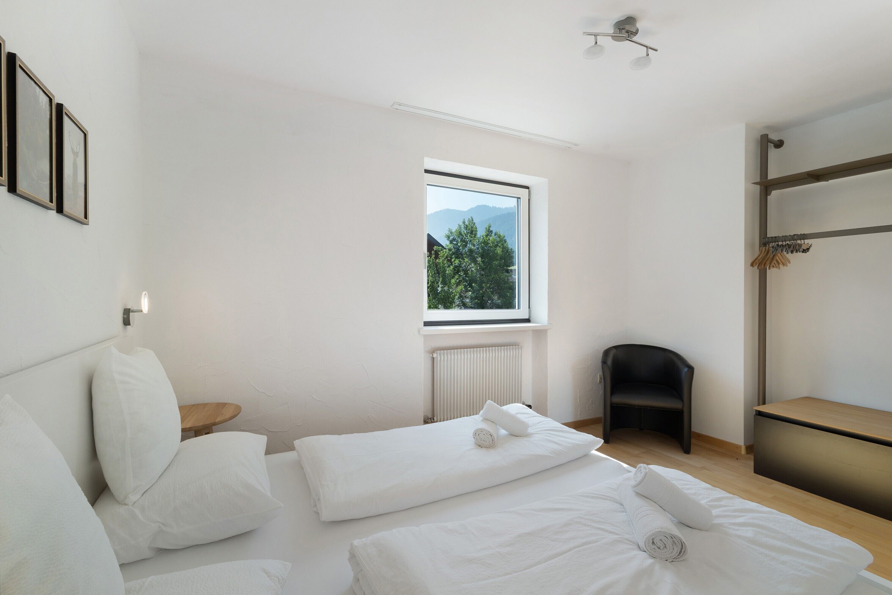Ein modernes Hotelzimmer mit großem Fenster, einem Bett mit weißer Bettwäsche, einem Holfnachtkästchen und einem schwarzen Sessel.