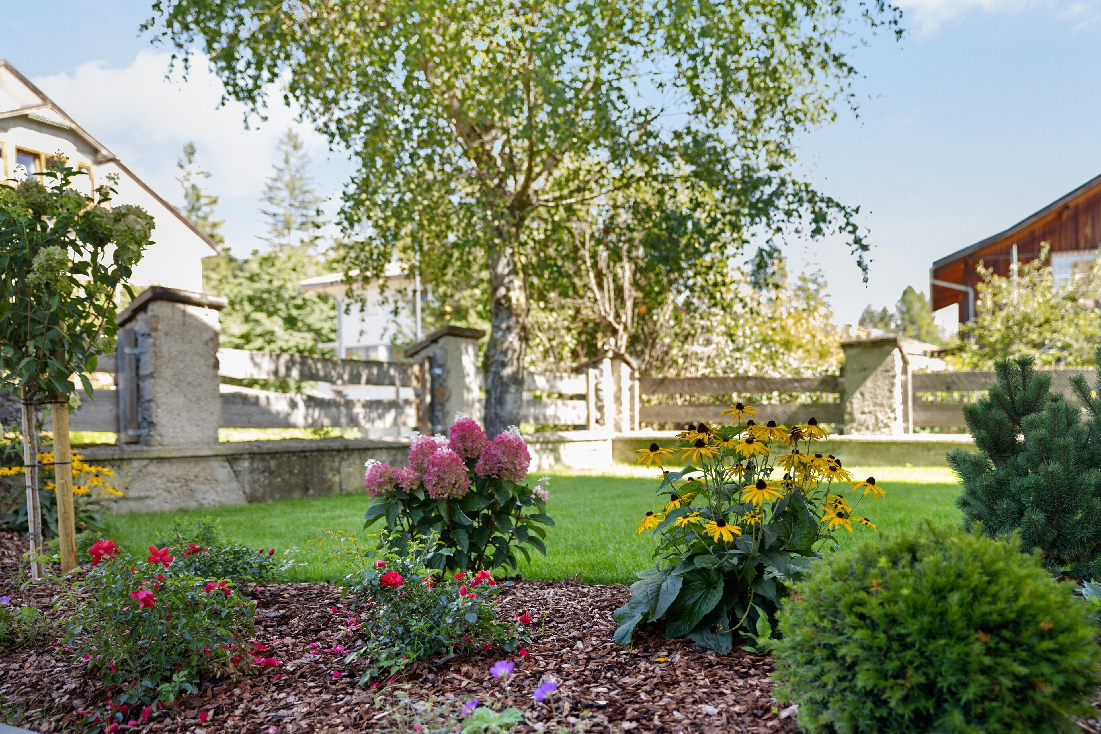 A garden with various plants and flowers, a stone wall, and a wooden building in the background.