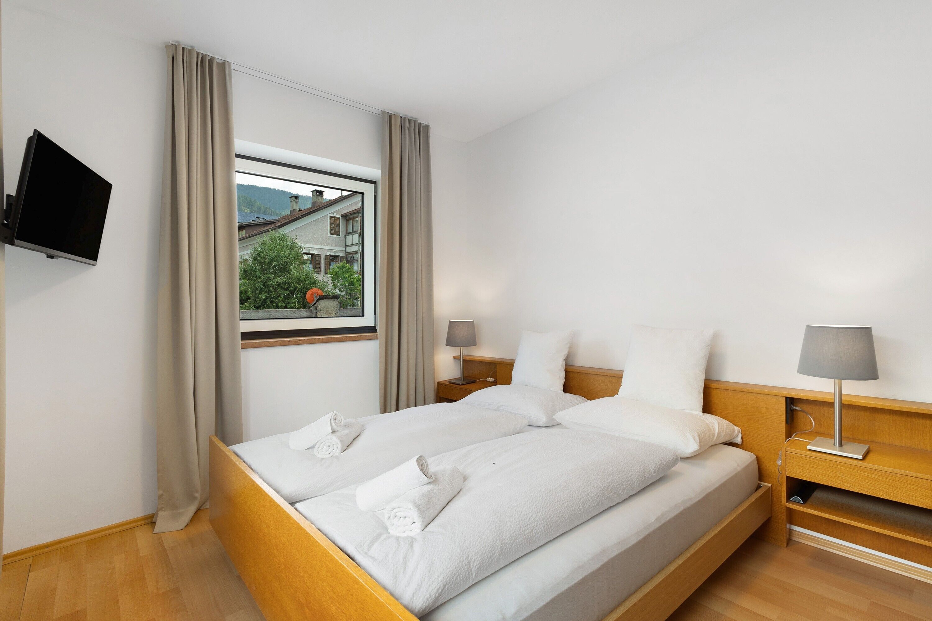 Habitación de hotel con cama, dos mesitas de noche, televisor montado en la pared, ventana con cortinas y vista a edificios.