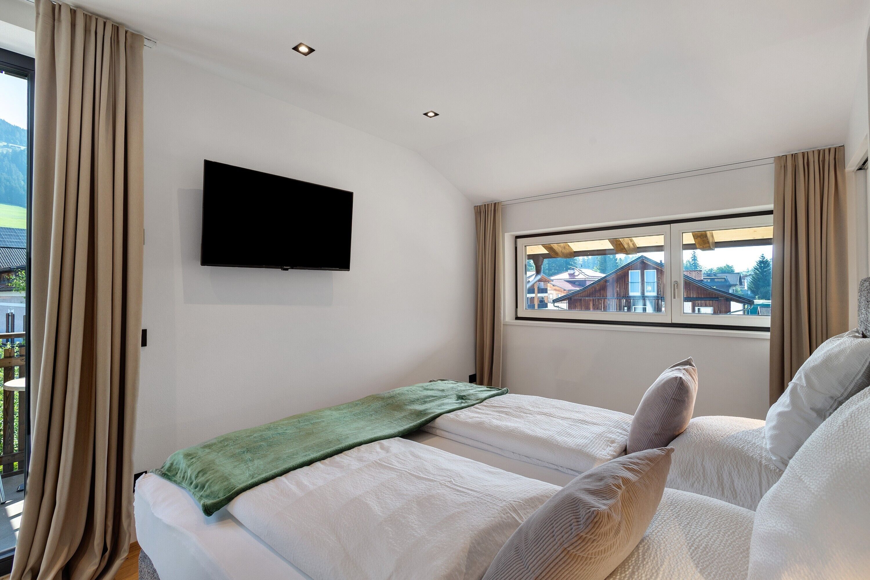 A bedroom with a bed, a television, a window with curtains, and a view of buildings.