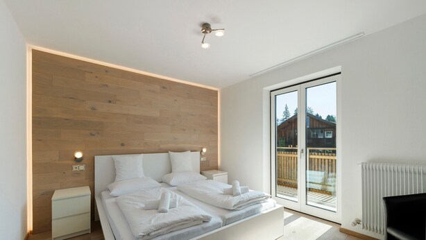 A modern bedroom with a wooden headboard, a bed with white linens, bedside lamps, and a sliding glass door to the outside. - Premium Apartment Hofer Elisabeth – Fast Internet, Free Parking (Niederdorf)