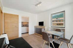 A modern room with a wooden door, a kitchenette, a dining area with a table and chairs, and a large window with a view of buildings.