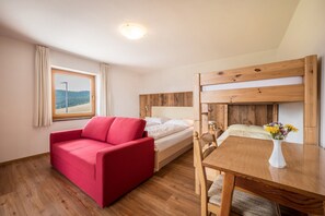 A room with a wooden bunk bed, a red sofa, a wooden dining table with chairs, and a window with curtains.