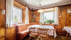 Un interior de madera con una mesa redonda, dos sillas, un banco y ventanas amplias con cortinas.