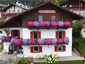 Un bâtiment à deux étages, avec une enseigne indiquant « Haus Adler », des balcons ornés de jardinières et un petit espace extérieur aménagé avec des sièges.