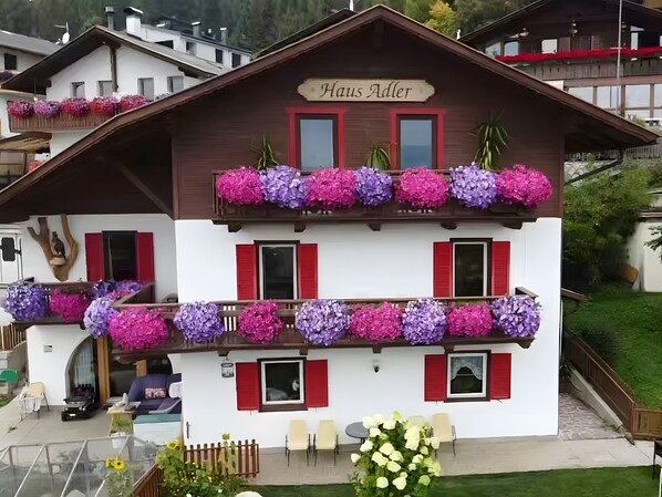 Un bâtiment à deux étages, avec une enseigne indiquant « Haus Adler », des balcons ornés de jardinières et un petit espace extérieur aménagé avec des sièges.