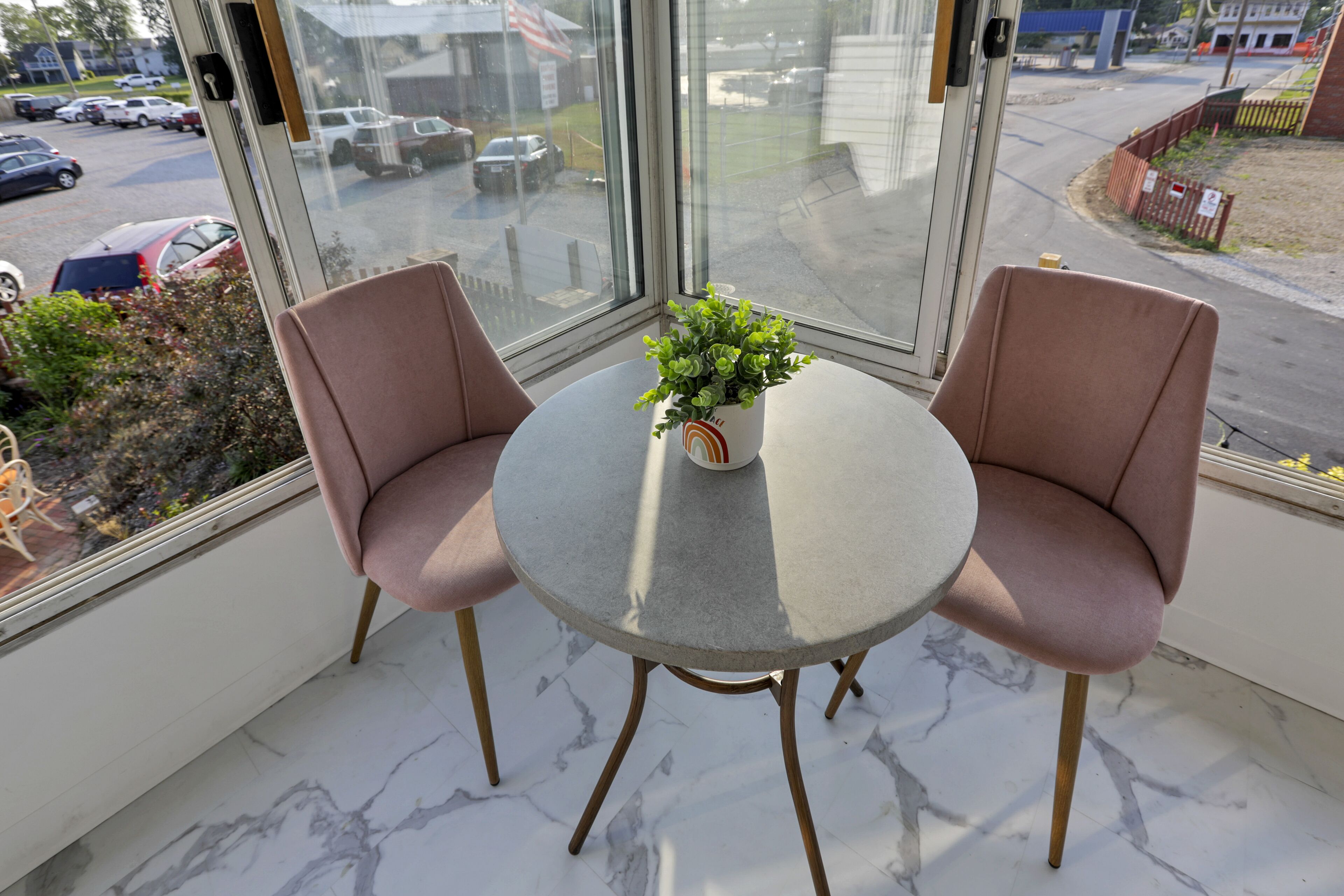 A room with two chairs and a table, a potted plant, and a view of a parking lot through large windows.