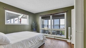 Een slaapkamer met een groot bed, een potplant, een raam met uitzicht op het water en een witte radiator.