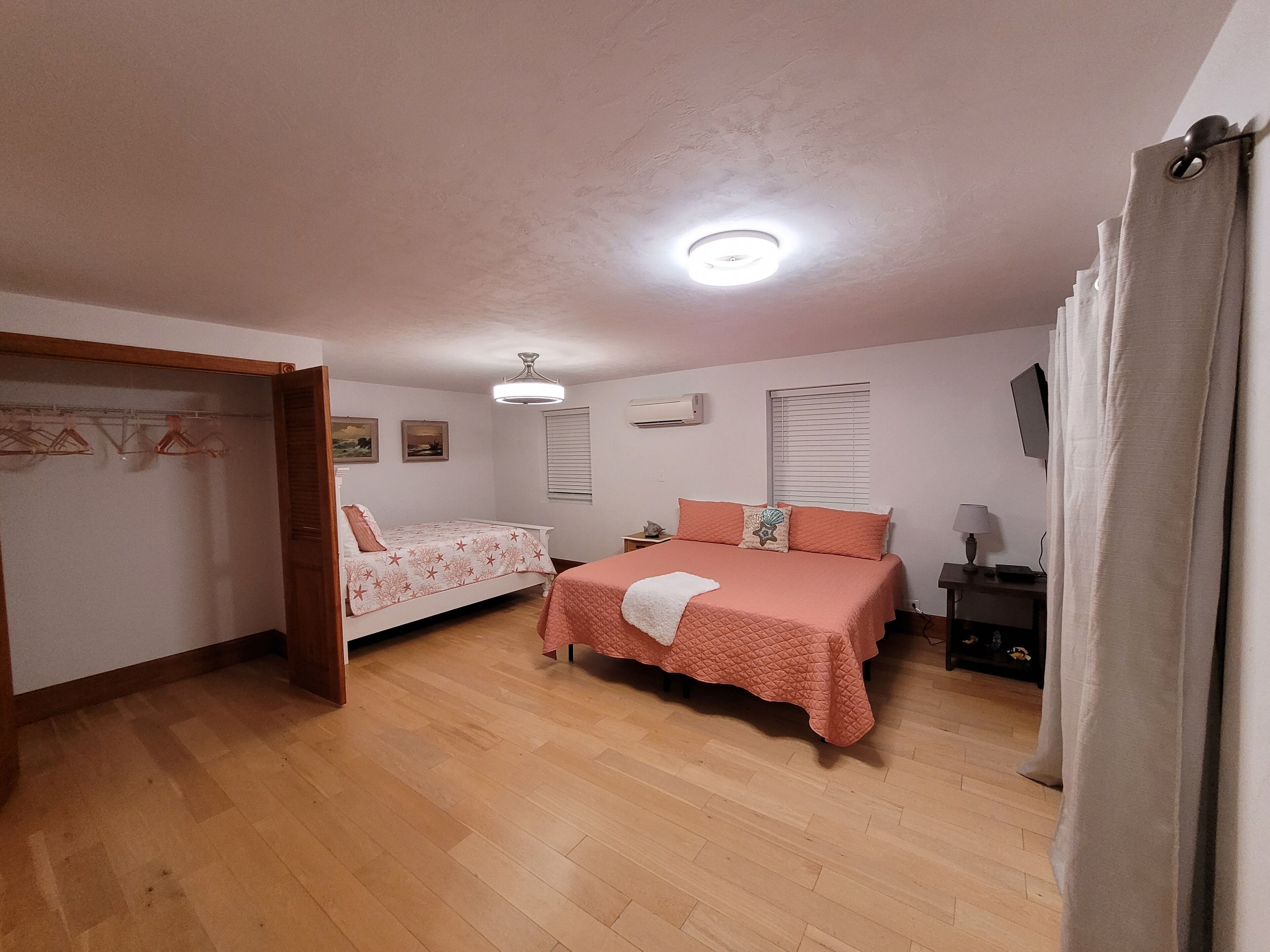 A bedroom with two beds, wooden flooring, a ceiling light, an air conditioning unit, and a TV on a stand.