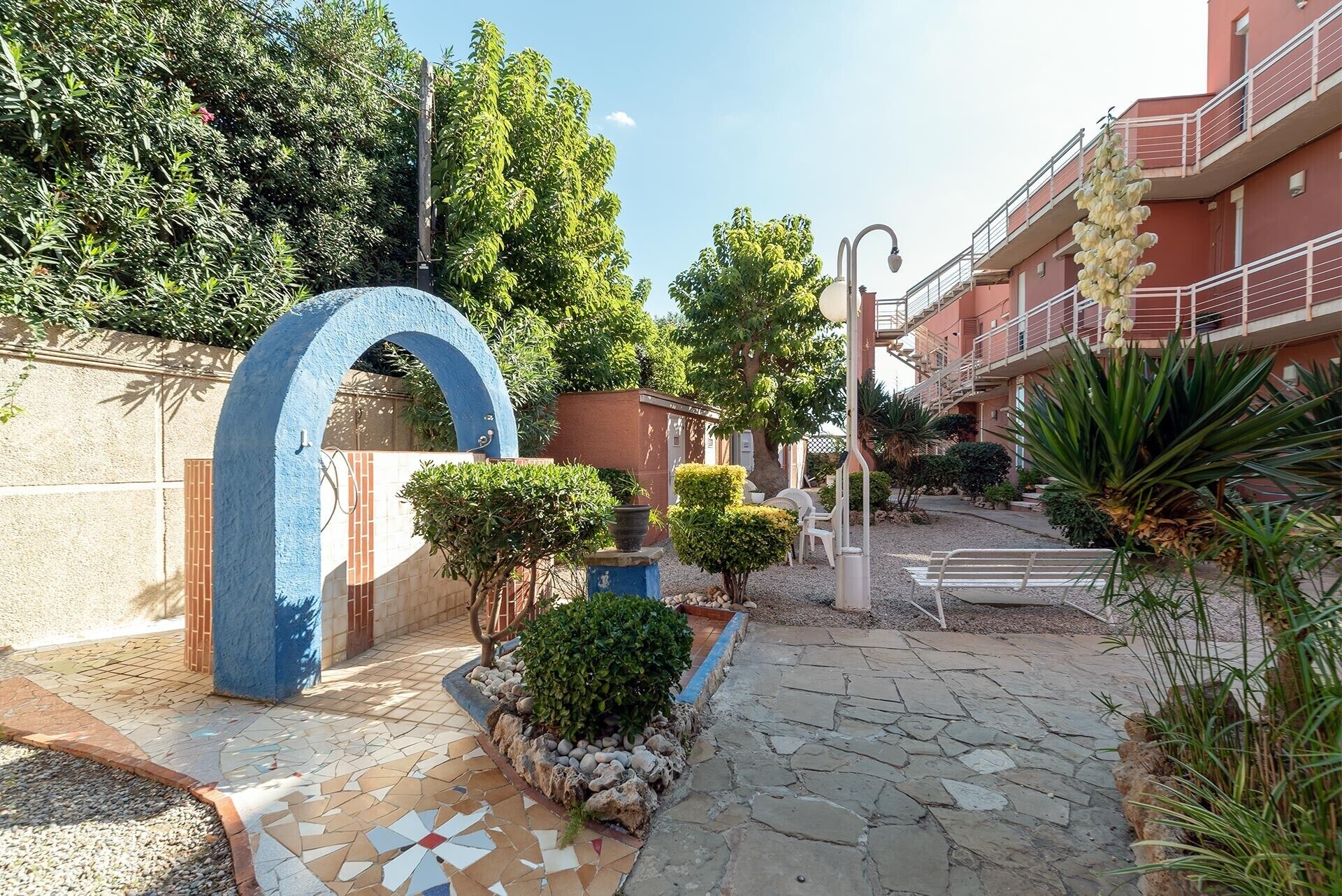 A paved pathway leading to a building entrance with a blue archway, surrounded by greenery and a stone-tiled area.