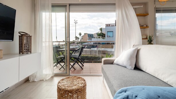 Ein modernes Wohnzimmer mit einer Couch, einem Fernseher, einem Balkon mit Tisch und Stühlen sowie einem Korbwäschekorb.