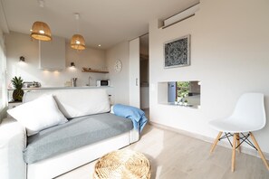 Un salon moderne comprenant un canapé, un fauteuil blanc, un panier en osier et un coin cuisine avec un four à micro-ondes.