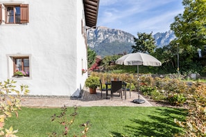 A white building with a patio, a table with chairs, a potted plant, and a mountain backdrop.