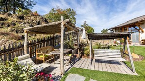 Eine Holzterrasse mit Gartenmöbeln, einer Pergola und einem Steinweg, der zu einem Gebäude mit Ziegeldach führt.