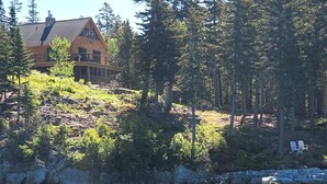 A log cabin with a wrap-around porch, situated on a rocky hillside with a view of a lake. - Sunset Cabin by the Sea: Log cabin w/ stunning ocean views. Unwind in paradise! (Roque Bluffs)