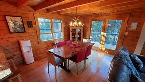 A wooden cabin interior with a dining table, chairs, a sofa, a water dispenser, and a chandelier. - Sunset Cabin by the Sea: Log cabin w/ stunning ocean views. Unwind in paradise! (Roque Bluffs)