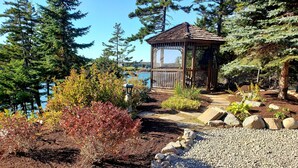 A gazebo with a wooden roof and open sides, surrounded by a gravel path, dense greenery, and a view of a lake. - Sunset Cabin by the Sea: Log cabin w/ stunning ocean views. Unwind in paradise! (Roque Bluffs)